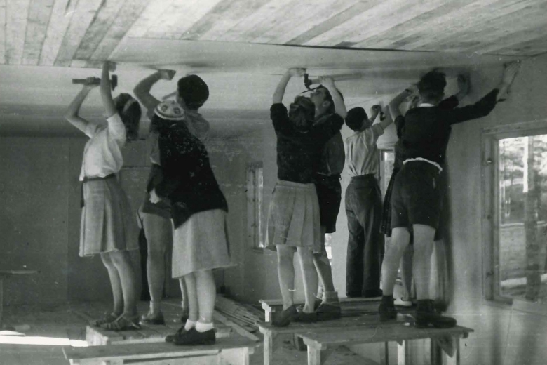 Eight young people standing on tables working on the ceiling of a building.