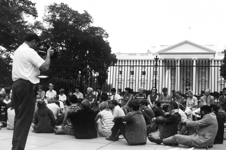 Protesters sit on the sidewalk in front of the White House while a man films.