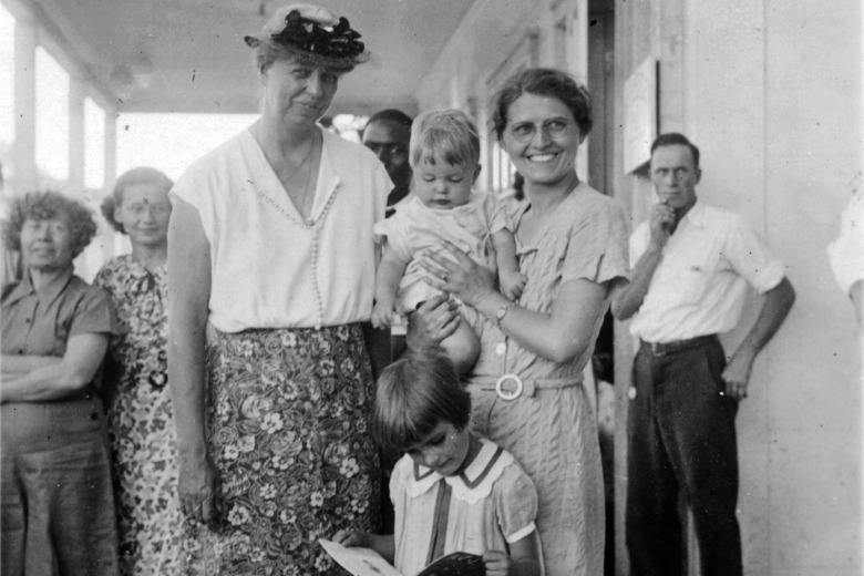 First Lady Eleanor Roosevelt standing beside a woman holding a baby and a young girl reading a book