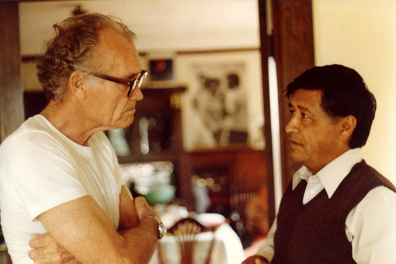 Two men standing, facing each other, speaking in a house.