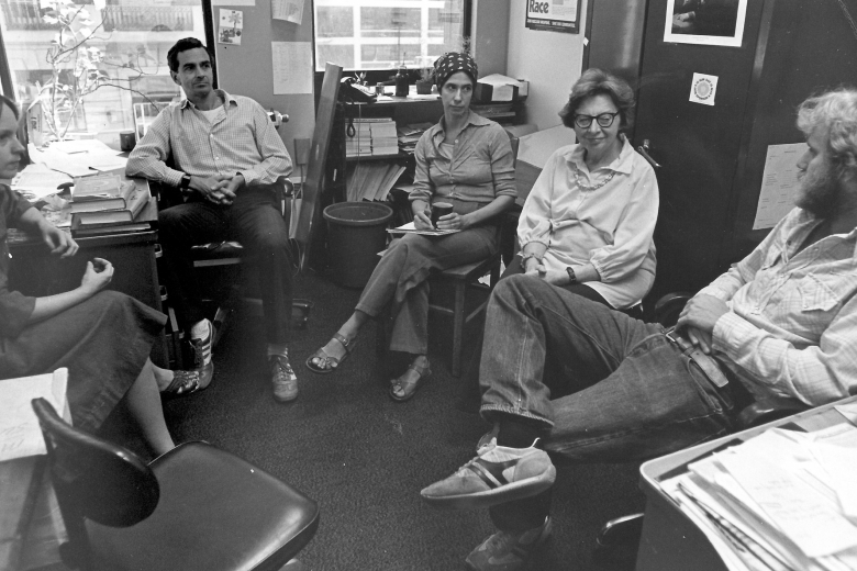 Five people sit in a circle in a small room full of files, talking to one another.