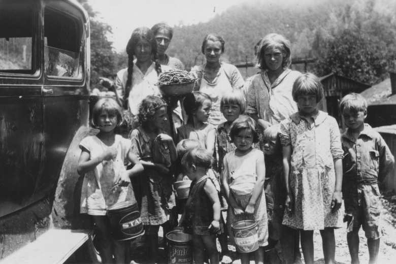 Group of women and children stand next to a car, holding pails.