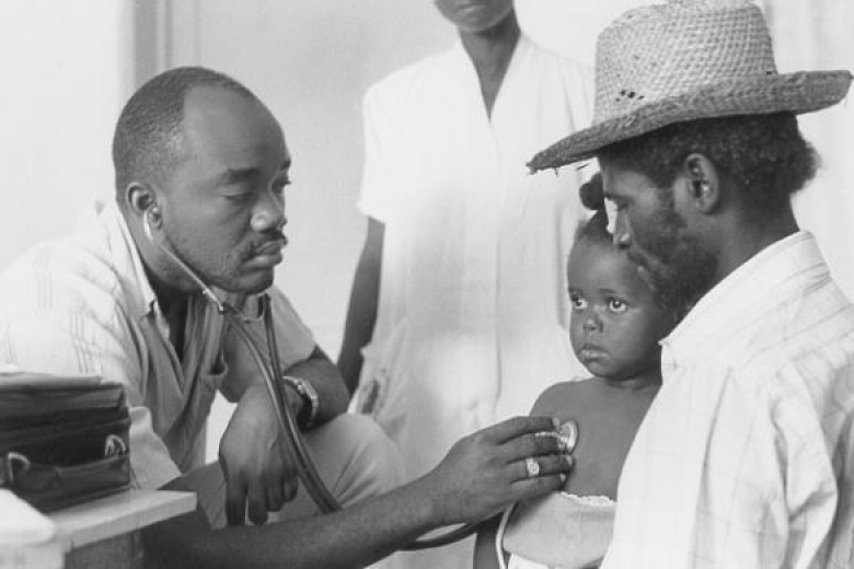 Child is examined by a doctor while a man wearing a hat holds the child on his lap.