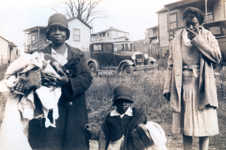 Two adult women and one child stand outside homes, holding clothing