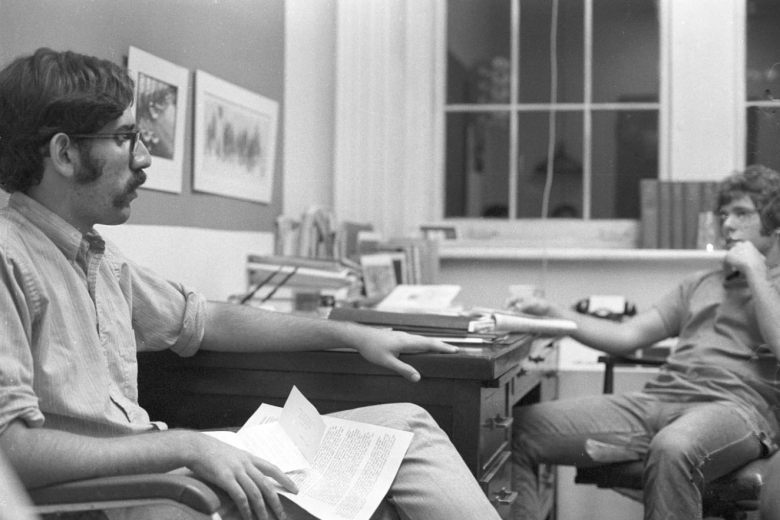 Two young men talk over a desk with papers on it.