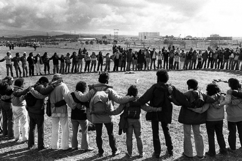 Circle of protesters with arms wrapped around one another, standing in a field.