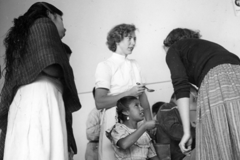 A young woman holding a spoon standing over a young girl, talking with two other young women.