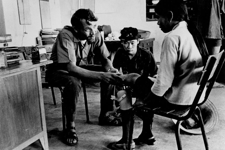 Young man speaks to a woman sitting in a chair with a prosthetic leg while another young man looks on.