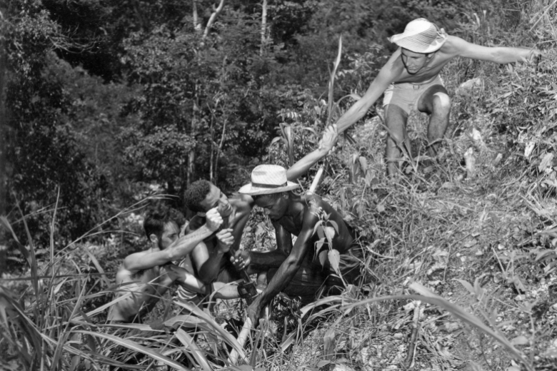 Four young men hold hands and crouch down to form a chain going down a hillside.