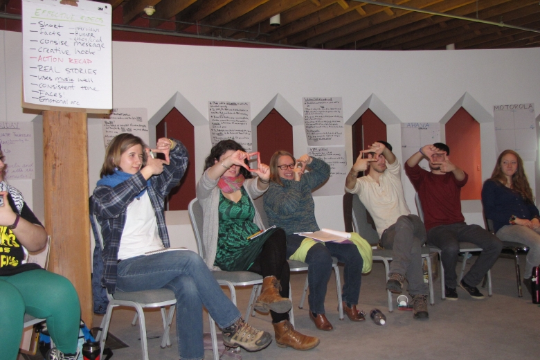 Six young adults sit in chairs and hold their hands up to their heads, forming a rectangle with their fingers.