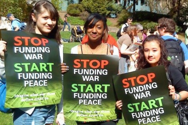 Three young women stand together, holding signs that read, "Stop funding war, start funding peace."