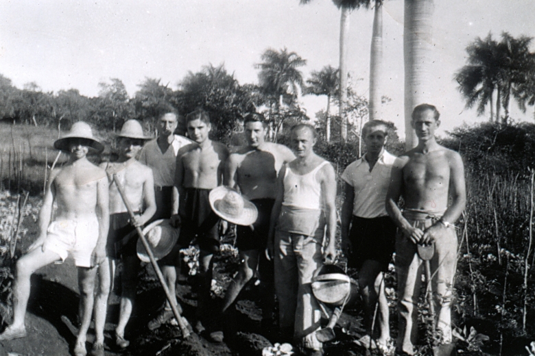 Men standing in front of palm trees, some wearing shirts, some without, some wearing wide-brimmed hats.