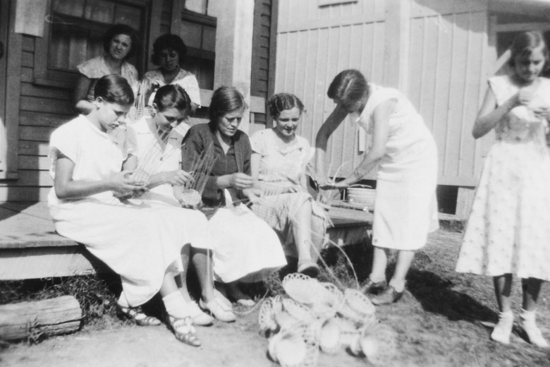 Women sitting on a porch helping each other make baskets