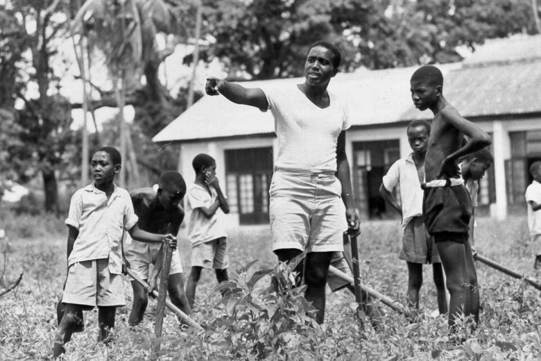 Young man in center of the group points to the left and speaks to group of five children working in a field.