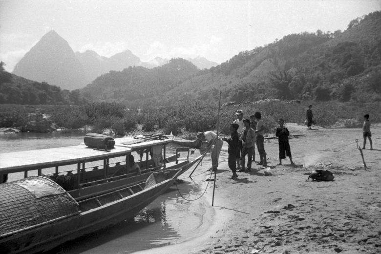 Small group of people stand together on a beach near a boat floating on the nearby body of water.