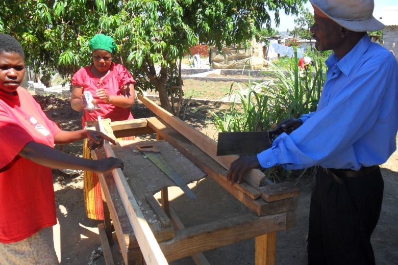 Group of three people do carpentry work in the shade near a tree.
