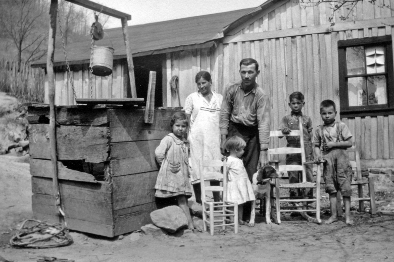 A family of 6 and a dog stand next to a well outside a house, with three wooden chairs