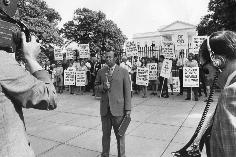 Man with microphone being filmed in front of a group of protesters holding signs with messages against the Vietnam War in front of the White House.