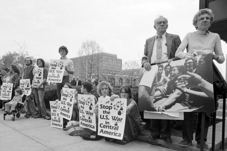 Group of people standing and sitting hold signs on a sidewalk.