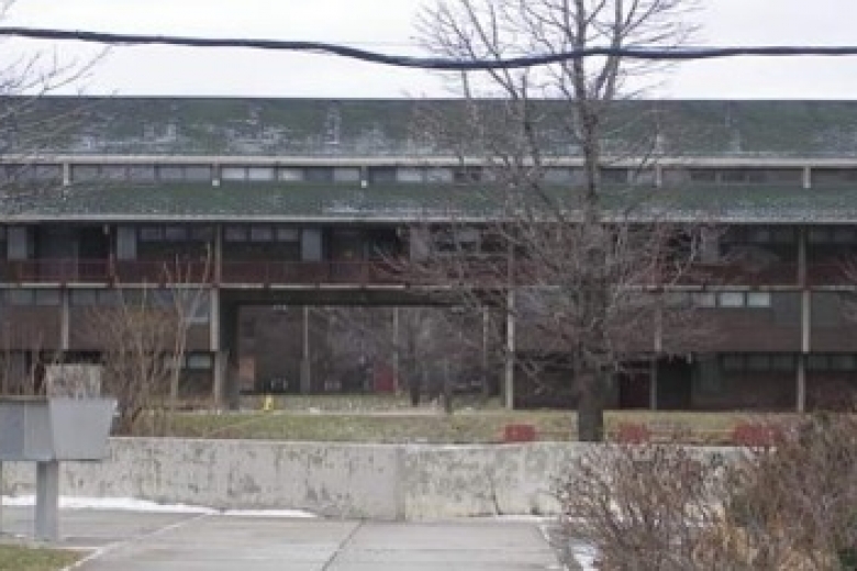 Old, grey building with empty parking lot in front of it.