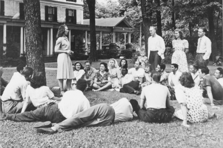 Group of young adults sits and stands together on a lawn, under a tree, with a building in the background.