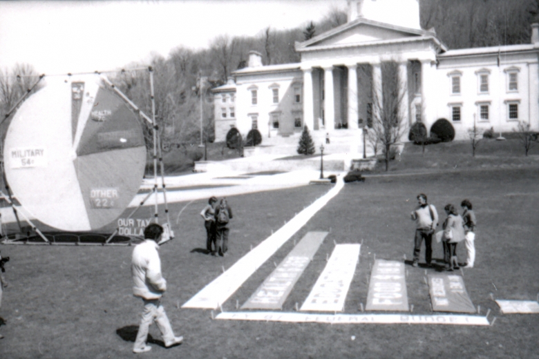 People examine a bar graph and pie chart displayed on the lawn in front of a state government building.