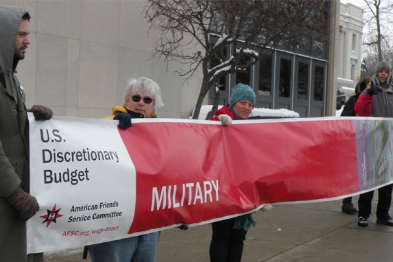 Group of people hold a large banner displaying the percentage of the U.S. discretionary budget spent on the military.