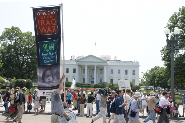 Group of people march together in front of the White House, one person holding a sign referencing the cost of the Iraq War.